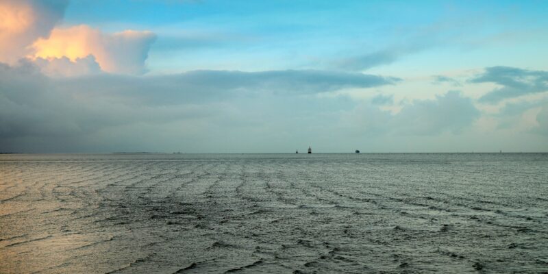 Blick über die Elbmündung bei Cuxhaven mit drei Frachtschiffen am Horizont, dramatischem Wolkenhimmel und warmem Lichtdurchbruch in der Abenddämmerung