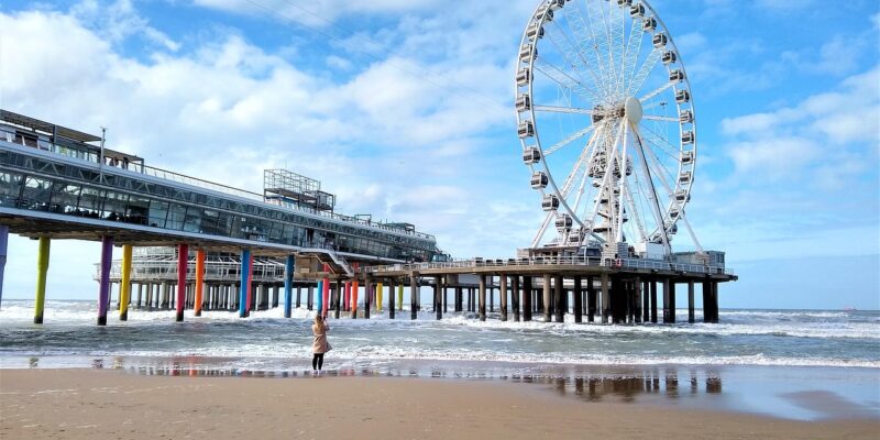 Scheveningen-Pier: Riesenrad am Strand (Tagesansicht) Die Pier von Scheveningen bei Den Haag mit dem SkyView-Riesenrad und den charakteristischen bunten Stützpfeilern, im Vordergrund der breite Sandstrand mit auflaufenden Wellen, eine Person fotografiert die Szene