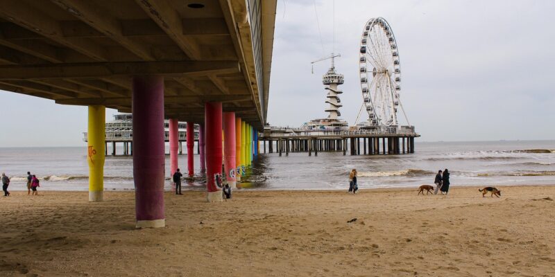 Bunte Stützpfeiler & Hund (Unteransicht) Blick unter die Pier von Scheveningen mit den bunten Stützpfeilern als Markenzeichen, im Vordergrund Spaziergänger und ein Hund am Strand, im Hintergrund Bungy-Tower und Riesenrad