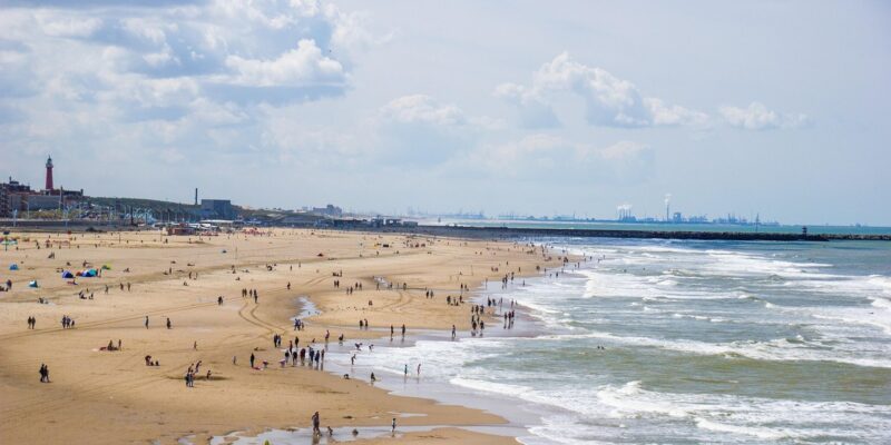 Scheveningen: Strand, Leuchtturm (Vuurtoren) & Besucher Der lange Sandstrand von Scheveningen an einem Sommertag mit zahlreichen Strandbesuchern, links der historische rote Vuurtoren-Leuchtturm und am Horizont die Hafensilhouette von Hoek van Holland