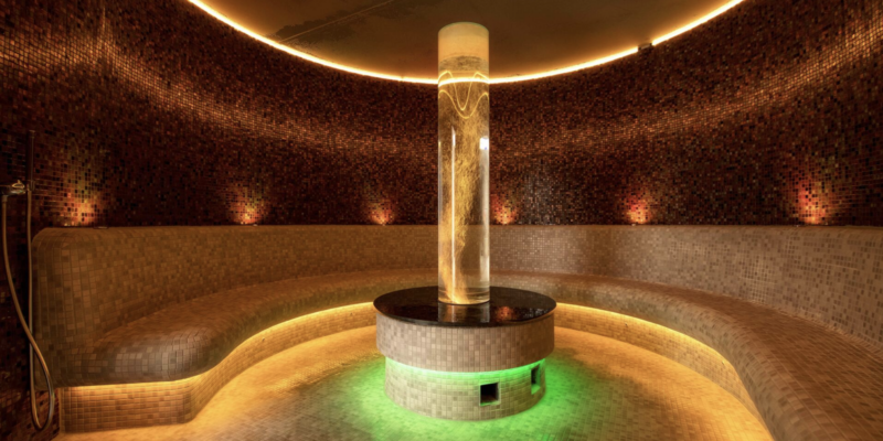 Das Dampfbad der Chiemgau Thermen – Wohlfühlen im sanften Nebel Das modern gestaltete, kreisrunde Dampfbad der Chiemgau Thermen mit warmen Mosaikfliesen und einer zentralen, beleuchteten Dampfsäule.
