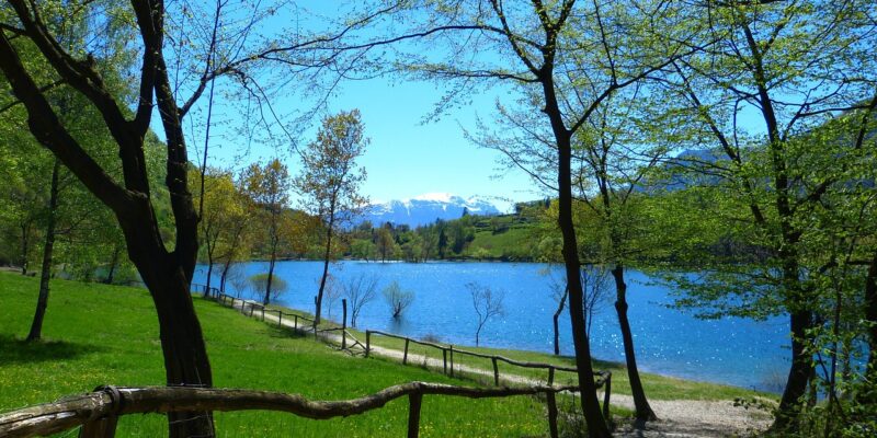 Uferweg am Lago di Tenno mit Bäumen, Holzgeländer, türkisblauem Wasser und schneebedeckten Bergen im Hintergrund