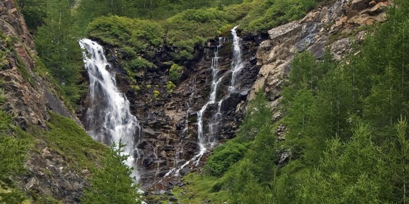Wasserfälle im Valle delle Cartiere fließen über dunkle Felsen, umgeben von grünen Bäumen und alpiner Landschaft