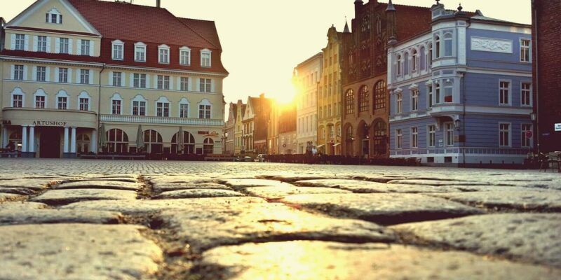 Eine Kopfsteinpflasterstraße in der Altstadt von Stralsund bietet bei Sonnenuntergang eine malerische Szenerie. Historische Gebäude mit unterschiedlicher Architektur säumen den Weg, darunter ein blaues und ein cremefarbenes Gebäude. Die Sonne geht zwischen den Gebäuden unter und wirft einen warmen, bernsteinfarbenen Schein über diese bezaubernde europäische Stadt.