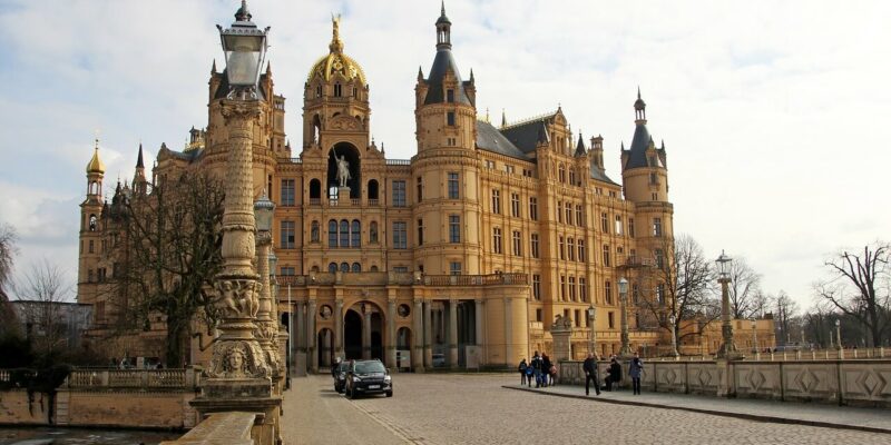 Besichtigen die Schätze des Schlosses Schwerin Ein großes, reich verziertes Schloss mit einer goldenen Kuppel und zahlreichen Türmchen vor einem wolkigen Himmel. Ein gepflasterter Weg führt zum Eingang. Menschen gehen spazieren und ein Auto parkt in der Nähe. Blattlose Bäume rahmen die Szene ein.