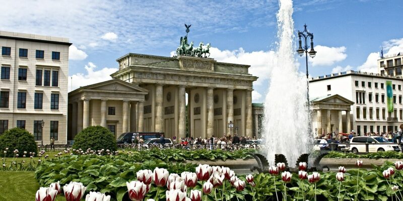 Blick auf das Tor Ein lebendiger Garten mit roten und weißen Tulpen im Vordergrund, einem großen Brunnen und dem historischen Brandenburger Tor in Berlin, mit einem blauen Himmel und vereinzelten Wolken darüber.