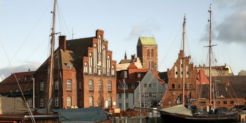 Ein malerischer Blick auf den Hafen der Hansestadt Wismar, mit traditionellen Backsteingebäuden im Hintergrund mit Stufengiebeln und einem hohen Kirchturm. Segelboote mit Masten liegen am Ufer vor Anker und spiegeln sich im ruhigen Wasser unter einem teilweise bewölkten Himmel.