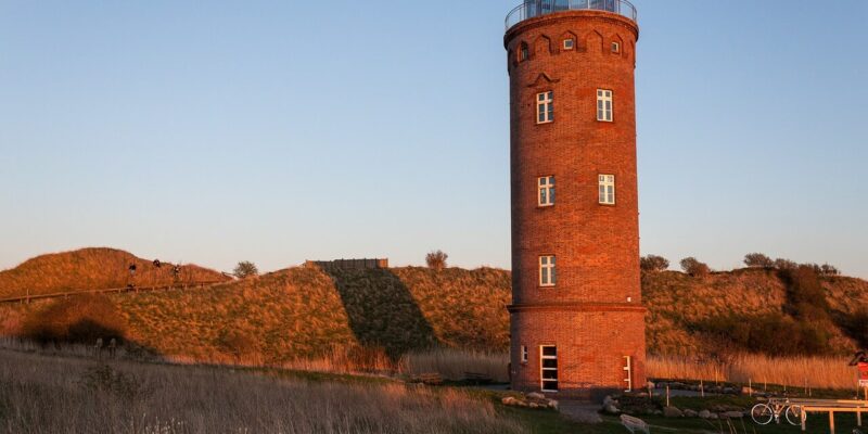 Ein hoher, zylindrischer Backsteinturm, der an Kap Arkona erinnert, mit mehreren Fenstern und einer Glaskuppel oben drauf, steht auf einer Rasenfläche. Die Sonne wirft warmes, goldenes Licht auf den Turm und die umgebende Landschaft. In der Nähe seines Fußes sind ein Pfad und ein paar Fahrräder zu sehen.