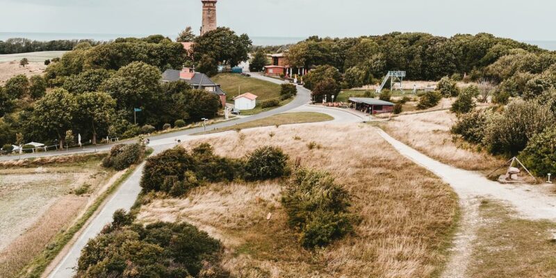 Eine malerische Ansicht einer ländlichen Landschaft mit einer kurvenreichen Straße, die zu einem roten Leuchtturm führt, der von Bäumen umgeben ist. Im Vordergrund sind goldene Felder zu sehen, während der Himmel bedeckt ist, was eine heitere Atmosphäre schafft.