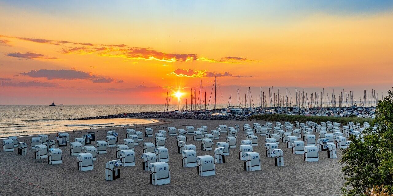 Ein malerischer Strand bei Sonnenuntergang mit Reihen weißer Stühle mit Blick aufs Meer fängt den Reiz der Sehenswürdigkeiten in Mecklenburg-Vorpommern ein. Der Himmel, eine leuchtende Mischung aus Orange und Lila, umrahmt Segelboote in der Nähe eines Wellenbrechers, während üppiges Grün den Sand säumt.
