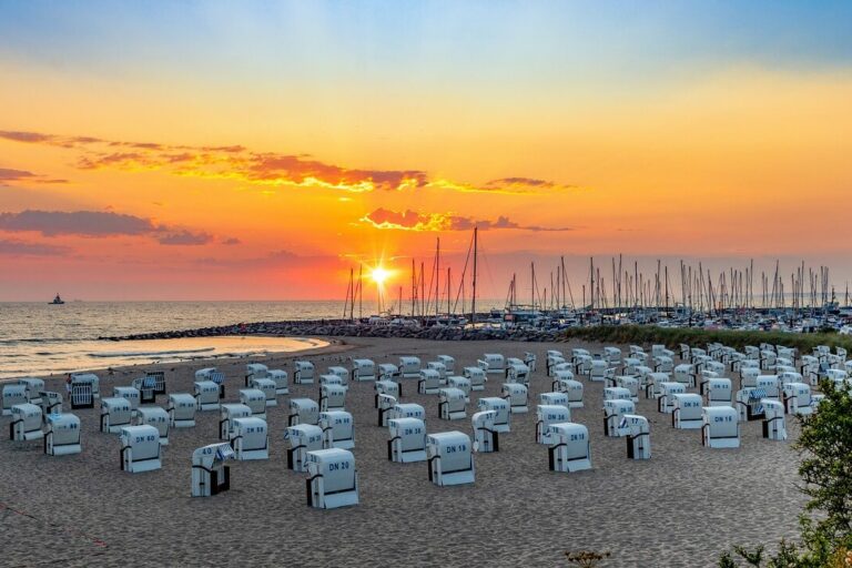 Ein malerischer Strand bei Sonnenuntergang mit Reihen weißer Stühle mit Blick aufs Meer fängt den Reiz der Sehenswürdigkeiten in Mecklenburg-Vorpommern ein. Der Himmel, eine leuchtende Mischung aus Orange und Lila, umrahmt Segelboote in der Nähe eines Wellenbrechers, während üppiges Grün den Sand säumt.
