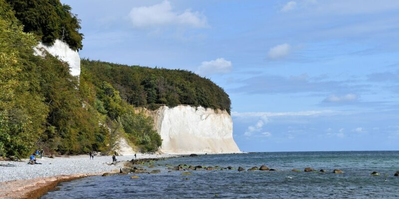 Der malerische Blick auf die felsige Küste im Nationalpark Jasmund bietet markante weiße Kreidefelsen und üppige grüne Bäume. Menschen schlendern am Ufer entlang, während sich das ruhige Meer unter einem teilweise bewölkten Himmel bis zum Horizont erstreckt.