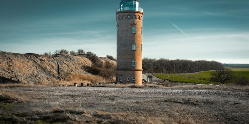 Ein hoher, zylindrischer Leuchtturm namens Peilturm Kap Arkona steht in einer kargen Landschaft mit spärlichem Gras. Der Himmel ist klar mit ein paar dünnen Wolken, und in der Ferne ist am Horizont eine Reihe von Bäumen zu erkennen.