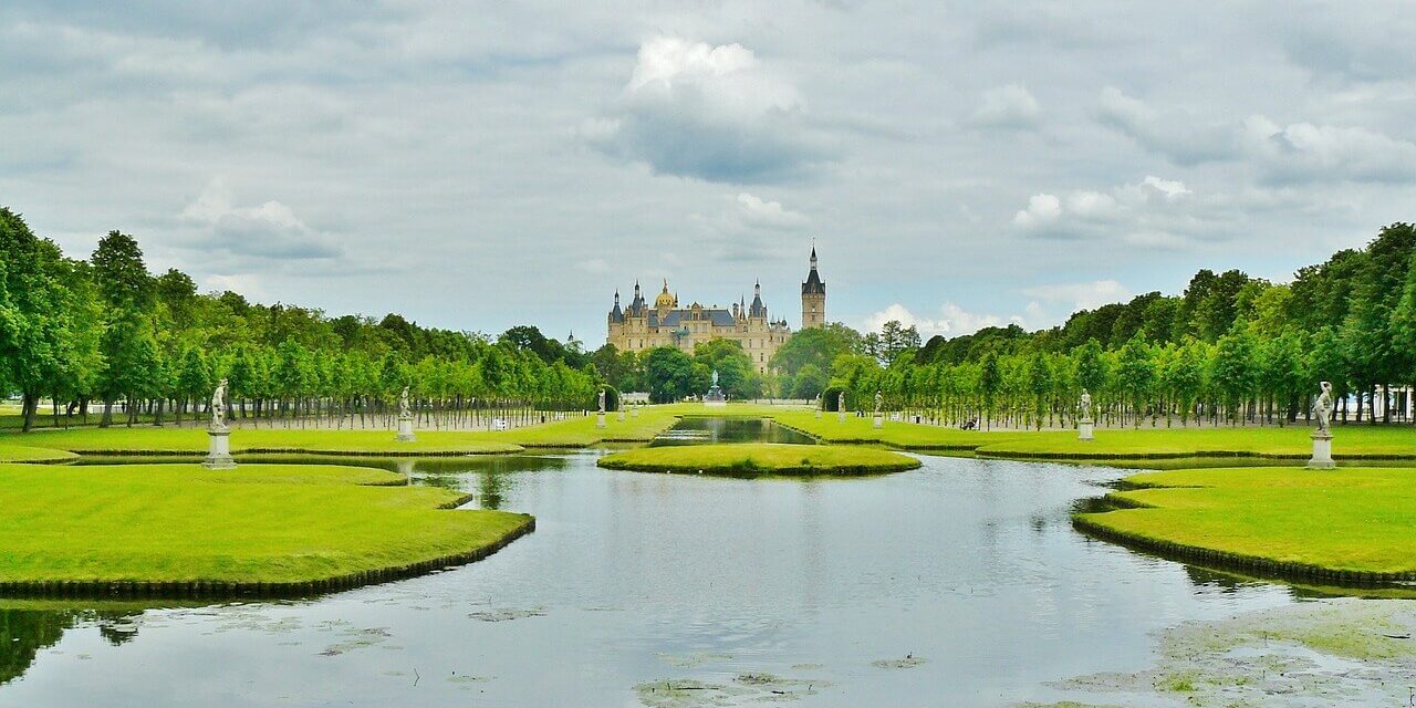 Schloss Schwerin Schloss Schwerin, ein prachtvolles Schloss mit spitzen Türmen, liegt in der Ferne, umgeben von üppigem Grün. Im Vordergrund spiegelt ein ruhiger Teich den bewölkten Himmel wider, gesäumt von gepflegten Rasenflächen und Baumreihen. Statuen schmücken beide Seiten des Wassers.