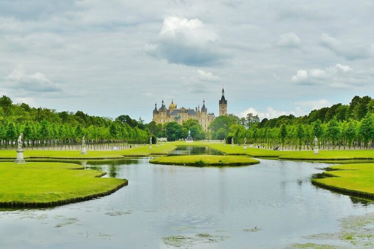 Schloss Schwerin Schloss Schwerin, ein prachtvolles Schloss mit spitzen Türmen, liegt in der Ferne, umgeben von üppigem Grün. Im Vordergrund spiegelt ein ruhiger Teich den bewölkten Himmel wider, gesäumt von gepflegten Rasenflächen und Baumreihen. Statuen schmücken beide Seiten des Wassers.