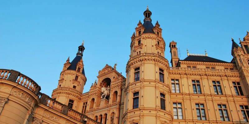 Eine Nahaufnahme des kunstvollen, historischen Schlosses Schwerin zeigt mehrere Türme und komplizierte architektonische Details vor einem klaren blauen Himmel. Die warme, sandfarbene Fassade und die dekorativen Elemente des Schlosses vermitteln ein Gefühl von Erhabenheit und zeitloser Eleganz.