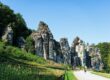 Eine Gruppe von Menschen geht auf die Externsteine zu, eines der schönsten Naturwunder Deutschlands, eine Reihe hoch aufragender Sandsteinfelsen, umgeben von üppigem Grün unter einem klaren blauen Himmel.