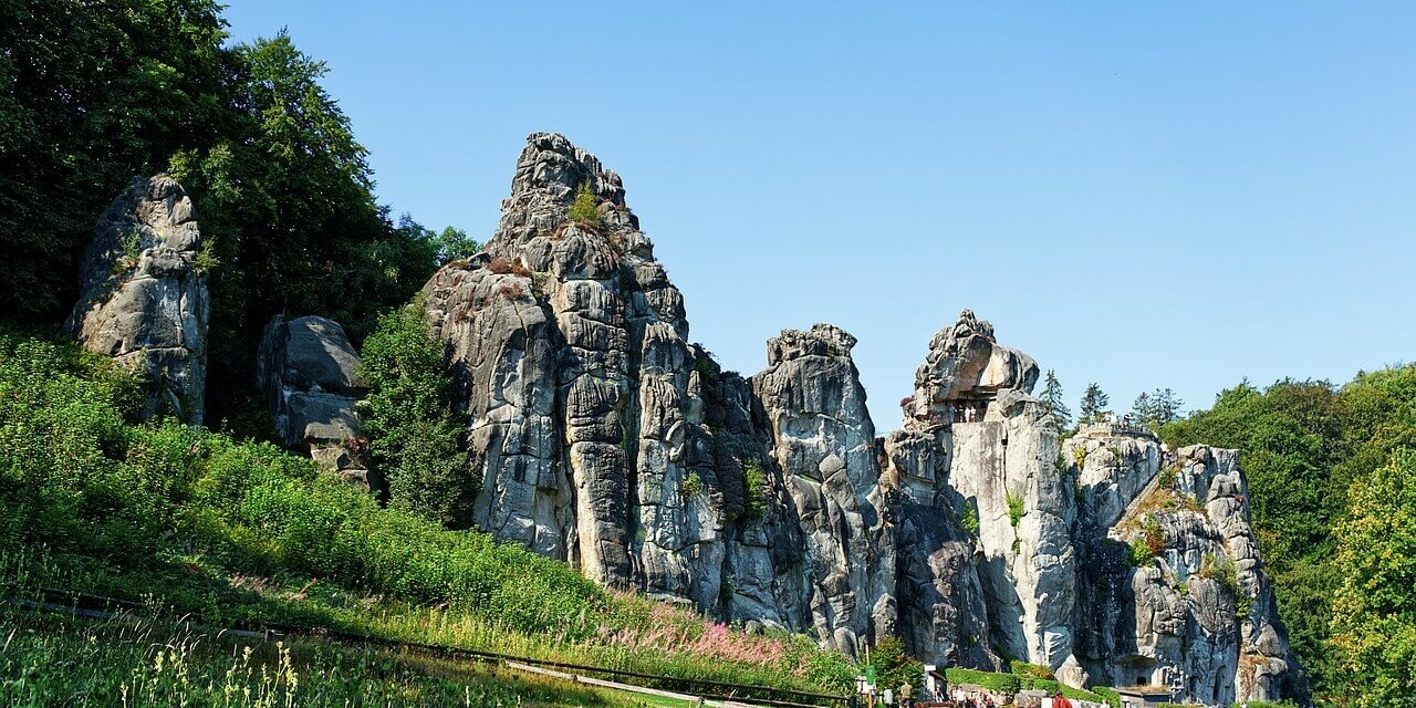 Eine Gruppe von Menschen geht auf die Externsteine zu, eines der schönsten Naturwunder Deutschlands, eine Reihe hoch aufragender Sandsteinfelsen, umgeben von üppigem Grün unter einem klaren blauen Himmel.