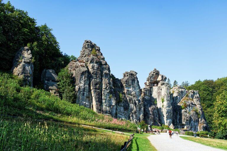 Eine Gruppe von Menschen geht auf die Externsteine zu, eines der schönsten Naturwunder Deutschlands, eine Reihe hoch aufragender Sandsteinfelsen, umgeben von üppigem Grün unter einem klaren blauen Himmel.