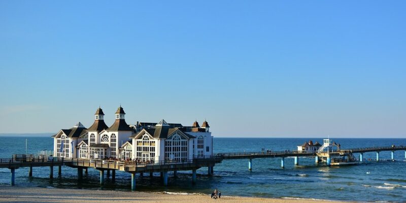 Eine malerische Anlegestelle, die an die Seebrücke Sellin erinnert, erstreckt sich in das ruhige blaue Meer und verfügt über ein charmantes Gebäude mit mehreren Spitzdächern. Am Sandstrand im Vordergrund sind einige Menschen zu sehen, die einen sonnigen Tag unter klarem Himmel genießen.