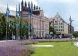 Eine lebhafte Straßenszene, typisch für Städte in Mecklenburg-Vorpommern, entfaltet sich mit einem Rasen und violetten Blumen im Vordergrund. Blaue Flaggen säumen die Straße, während bunte Gebäude, darunter ein rosafarbenes mit Bogenfenstern, Charme verleihen. Menschen schlendern unter dem bewölkten Himmel.