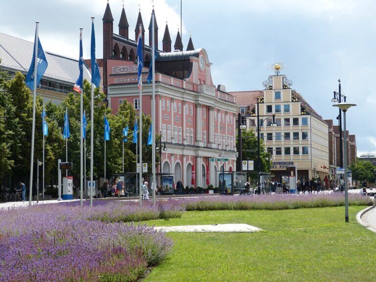 Eine lebhafte Straßenszene, typisch für Städte in Mecklenburg-Vorpommern, entfaltet sich mit einem Rasen und violetten Blumen im Vordergrund. Blaue Flaggen säumen die Straße, während bunte Gebäude, darunter ein rosafarbenes mit Bogenfenstern, Charme verleihen. Menschen schlendern unter dem bewölkten Himmel.