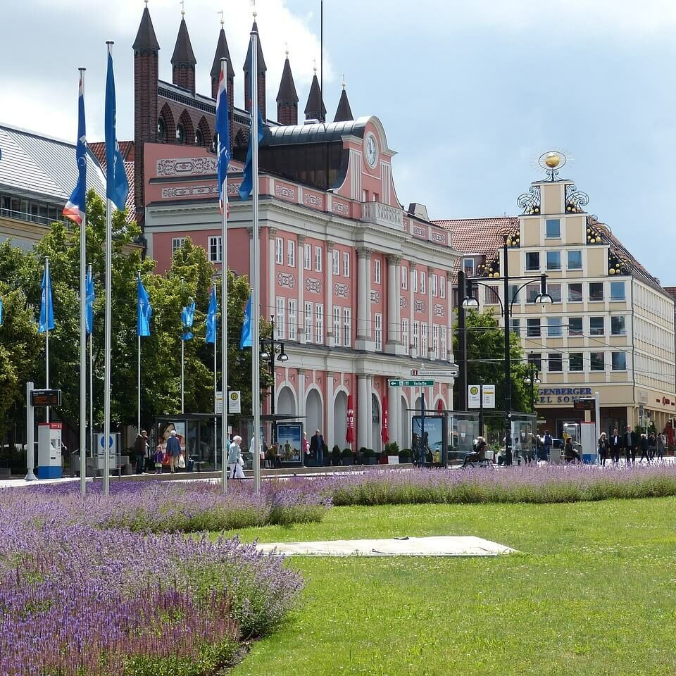 Eine lebhafte Straßenszene, typisch für Städte in Mecklenburg-Vorpommern, entfaltet sich mit einem Rasen und violetten Blumen im Vordergrund. Blaue Flaggen säumen die Straße, während bunte Gebäude, darunter ein rosafarbenes mit Bogenfenstern, Charme verleihen. Menschen schlendern unter dem bewölkten Himmel.