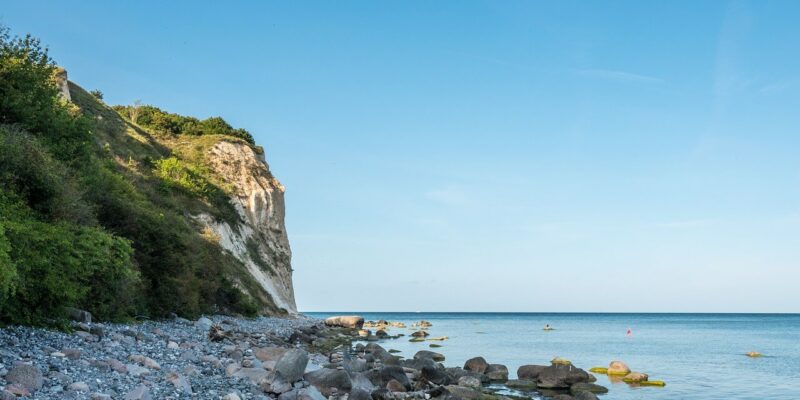 Eine Küstenszene an der Steilküste Kap Arkona auf der Insel Rügen zeigt einen felsigen Strand mit verstreuten Steinen, der zu einer steilen, grasbewachsenen Klippe auf der linken Seite hinaufführt. Das ruhige blaue Meer erstreckt sich unter einem klaren blauen Himmel bis zum Horizont.
