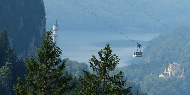 Eine malerische Aussicht auf eine Seilbahn, die über eine bewaldete Landschaft mit einem hohen Turm, einem Schloss in der Ferne und einem ruhigen See, umgeben von nebligen Bergen, fährt. Im Vordergrund sind hohe immergrüne Bäume zu sehen.