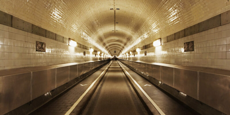 Alte Elbtunnel Der Alte Elbtunnel in Hamburg besteht aus einem langen, symmetrischen Durchgang mit weiß gefliesten Wänden und einer Decke, die von Lichtreihen beleuchtet wird. Die leere Straße im Inneren zeigt sichtbare Fahrspuren und schwache Reflexionen, während der Tunnel in die Ferne verschwindet und einen faszinierenden Fluchtpunkteffekt erzeugt.