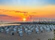 Ein Strand bei Sonnenuntergang in Mecklenburg-Vorpommern besticht durch Reihen weißer Stühle mit Blick auf das Meer. Der Himmel strahlt in Orange-, Rosa- und Lilatönen, während in der Nähe angedockte Segelboote eine ruhige Szenerie schaffen. Perfekte Ausflugsziele für alle, die sanft am Horizont versinkende Sonnenuntergänge zu schätzen wissen.