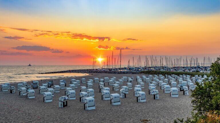 Ein Strand bei Sonnenuntergang in Mecklenburg-Vorpommern besticht durch Reihen weißer Stühle mit Blick auf das Meer. Der Himmel strahlt in Orange-, Rosa- und Lilatönen, während in der Nähe angedockte Segelboote eine ruhige Szenerie schaffen. Perfekte Ausflugsziele für alle, die sanft am Horizont versinkende Sonnenuntergänge zu schätzen wissen.