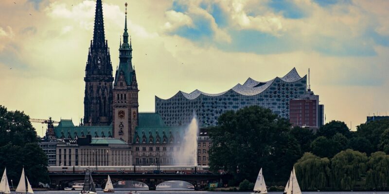 Außenalster Ein Panoramablick auf eine Stadtlandschaft mit einer großen Kirche und einem modernen Konzertsaal unter einem bewölkten Himmel. Segelboote gleiten anmutig auf der Außenalster, umgeben von üppigem Grün, was der lebhaften Szenerie Charme verleiht.