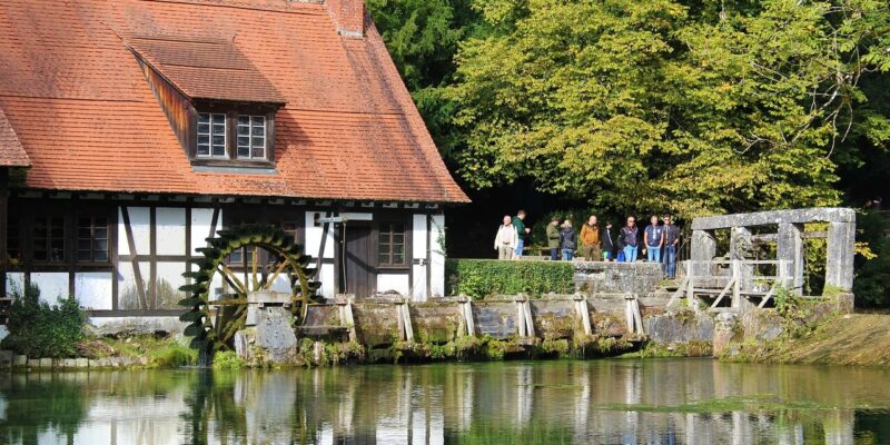 Eine historische Fachwerkmühle mit Wasserrad ziert den Rand des Blautopfs in Blaubeuren, ihr Spiegelbild spiegelt sich im ruhigen Teich. In der Nähe steht eine Gruppe auf einem Holzsteg und genießt die ruhige Schönheit umgeben von üppigen Bäumen.