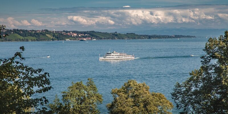 Eine malerische Aussicht auf den Bodensee mit einer Fähre in der Mitte, umgeben von üppigen grünen Bäumen im Vordergrund. Die entfernte Uferlinie zeichnet sich durch sanfte Hügel und einige Gebäude unter einem teilweise bewölkten Himmel aus.