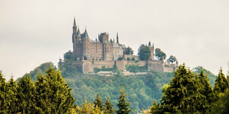 Ein majestätisches Schloss, das an die Burg Hohenzollern erinnert, thront auf einem üppigen, bewaldeten Hügel, umgeben von dichtem Grün. Der Himmel ist bedeckt und bietet eine dramatische Kulisse für die historische Architektur mit ihren zahlreichen Türmen und Steinmauern.