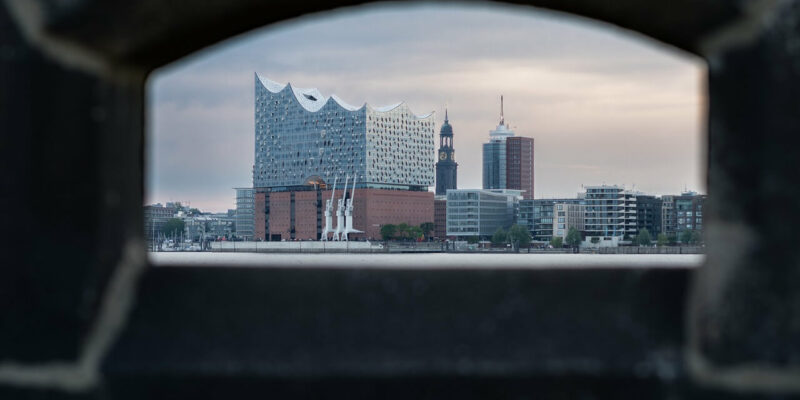 Elbphilharmonie Werfen Sie einen Blick auf die Elbphilharmonie in Hamburg, perfekt eingerahmt durch eine Steinöffnung. Die moderne Glasfassade bildet einen schönen Kontrast zu den historischen Gebäuden unter einem stimmungsvollen, wolkigen Himmel.