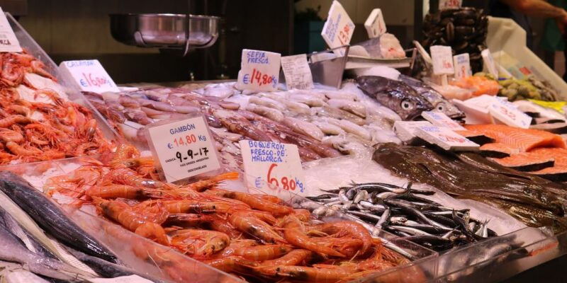 Fischmarkt Eine Fischmarkt-Auslage auf dem Hamburger Fischmarkt bietet eine Auswahl an frischem Fisch und Schalentieren auf Eis. Zu sehen sind Garnelen, Lachs und verschiedene Fischarten, mit Preisschildern in Euro. Im Hintergrund bietet ein geschäftiger Fischstand noch mehr Meeresspezialitäten an.