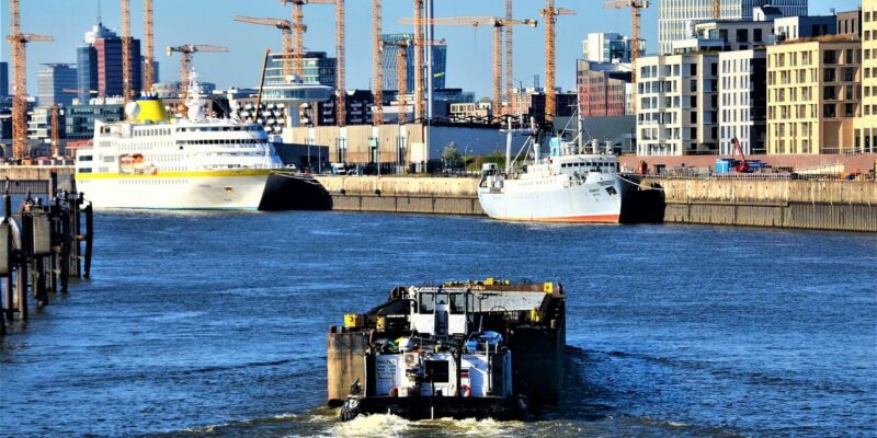 HafenCity Ein Frachtschiff fährt auf einem Fluss auf zwei große angedockte Schiffe zu. Im Hintergrund ist die Skyline der HafenCity in Hamburg mit zahlreichen Baukränen und modernen Gebäuden gesäumt, die sich unter einem klaren blauen Himmel am Wasser entlang erstrecken.