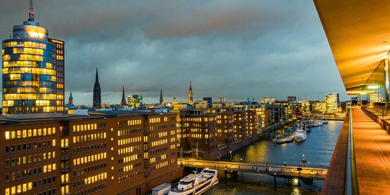 Hamburg Sehenswürdigkeiten Eine Stadtlandschaft in der Abenddämmerung mit beleuchteten Bürogebäuden entlang eines Kanals zeigt einige der Sehenswürdigkeiten Hamburgs. Boote liegen entlang des Wasserwegs vor Anker und eine Brücke überquert den Kanal. Der bewölkte Himmel fängt einen warmen Schein der Lichter der Stadt ein, die sich im Wasser spiegeln.