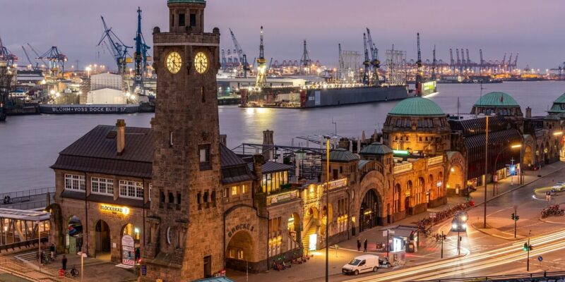Hamburger Hafen Ein ruhiger Abendblick auf die St. Pauli Landungsbrücken in Hamburg, Deutschland. Der Uhrenturm ist auffällig beleuchtet, die umliegenden Gebäude sind ebenfalls beleuchtet. Vor dem Abendhimmel bilden der Hamburger Hafen und die Kräne eine beeindruckende Kulisse, während Autos und Straßenlaternen die Straßen säumen.