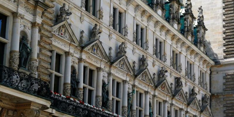 Hamburger Rathaus Das Hamburger Rathaus besticht durch eine detailreiche Fassade mit Reihen verzierter Fenster und Statuen in Nischen im klassischen Stil. Seine Architektur umfasst aufwendige Steinmetzarbeiten und ein dekoratives schwarzes Geländer, das mit kleinen Pflanzen geschmückt ist und die Erhabenheit dieses historischen Gebäudes widerspiegelt.