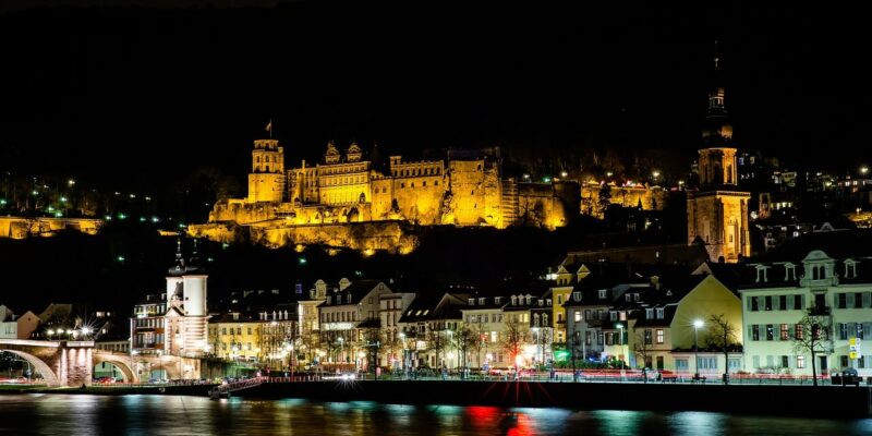 Nachtansicht von Heidelberg, Deutschland, mit dem beleuchteten Heidelberger Schloss auf einem Hügel. Der Neckar glitzert, während er die bunten Lichter der Gebäude am Wasser reflektiert. Links erhebt sich die Alte Brücke anmutig vor dem Nachthimmel.