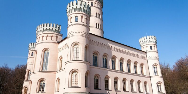 Das majestätische Jagdschloss Granitz, ein großes rosa Schloss mit runden Türmen und gewölbten Fenstern, erhebt sich stolz vor einem klaren blauen Himmel. Besucher schlendern in der Nähe umher, während ein paar Fahrzeuge davor parken. Umgeben von üppigen Bäumen fängt dieser historische Ort die Essenz zeitloser Eleganz ein.