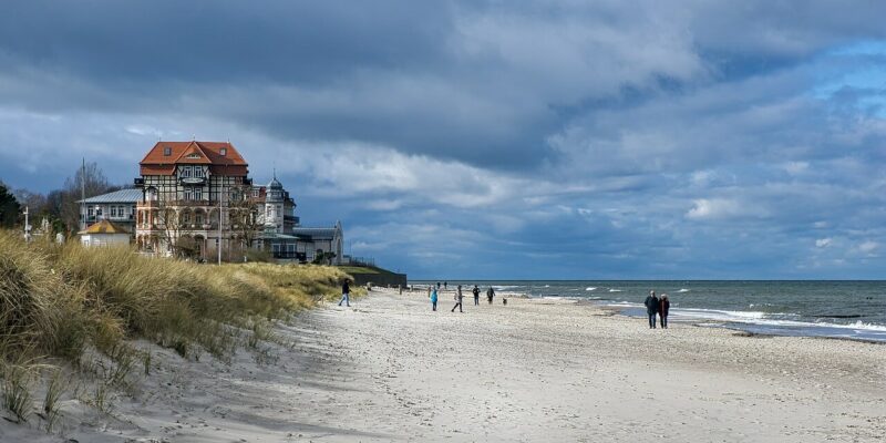 Kühlungsborn Strand Ein Sandstrand am Kühlungsborner Strand mit ein paar Leuten, die unter einem bewölkten Himmel am Ufer entlangspazieren. Links steht ein großes, reich verziertes Gebäude mit roten Dächern nahe dem Rand des Strandes, umgeben von grasbewachsenen Dünen. Das Meer ist ruhig mit sanften Wellen.