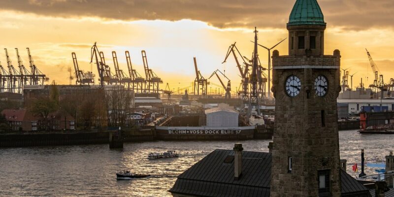 Landungsbrücken Eine Szene am Flussufer bei Sonnenuntergang mit dem ikonischen Uhrturm der Landungsbrücken im Vordergrund. Im Hintergrund sind mehrere Kräne auf einer Werft unter einem warmen, leuchtenden Himmel zu sehen. Das Wasser spiegelt die goldenen Töne des Sonnenuntergangs wunderschön wider.