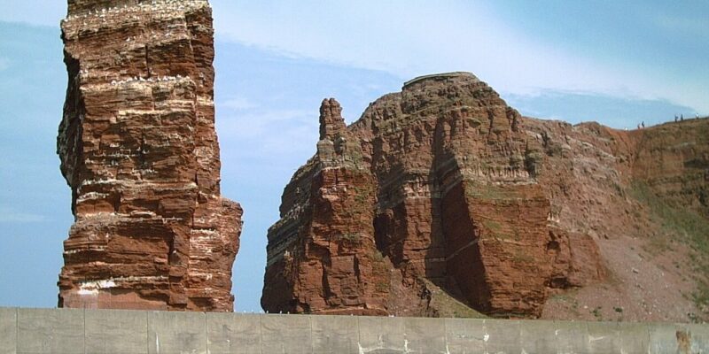 Hohe rote Felsformationen, die an die Lange Anna auf Helgoland erinnern, erheben sich hinter einer Betonmauer am Wasserrand vor einem blauen Himmel. Möwen fliegen über den Klippen und bereichern die zerklüftete Küstenlandschaft.