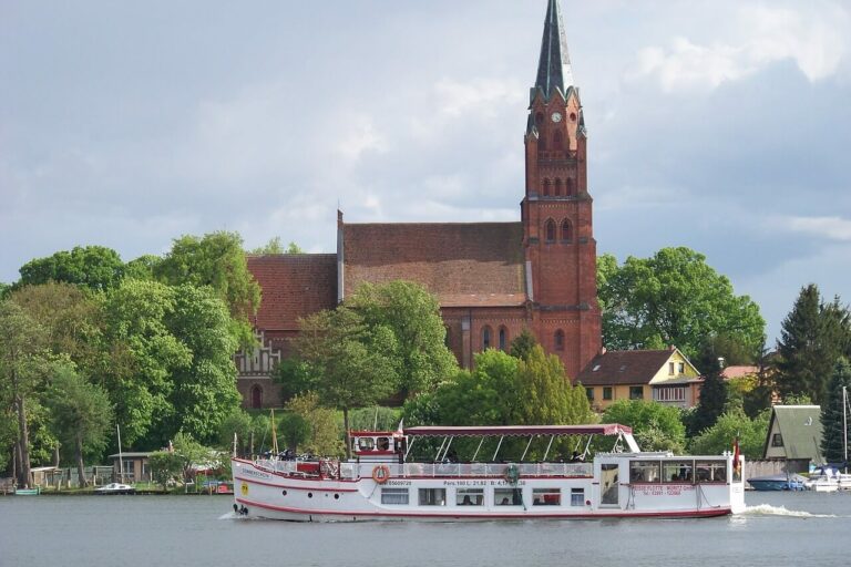 Mecklenburgischen Seenplatte Ein weißes Ausflugsboot mit Passagieren fährt auf einem Fluss in der Mecklenburgischen Seenplatte und passiert eine historische rote Backsteinkirche mit einem hohen Kirchturm. Die Kirche ist elegant von üppigen grünen Bäumen unter einem wolkigen Himmel eingerahmt.