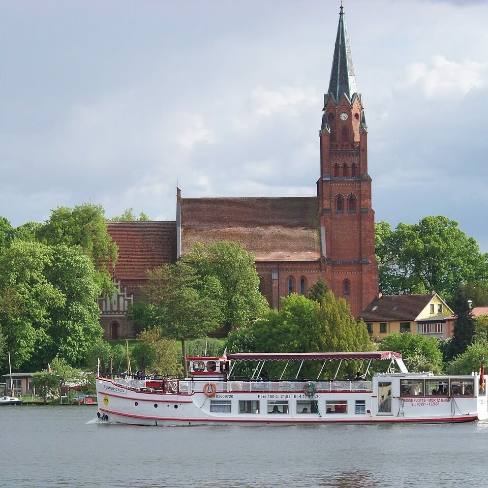Ein weißes Ausflugsboot mit Passagieren fährt auf einem Fluss in der Mecklenburgischen Seenplatte und passiert eine historische rote Backsteinkirche mit einem hohen Kirchturm. Die Kirche ist elegant von üppigen grünen Bäumen unter einem wolkigen Himmel eingerahmt.