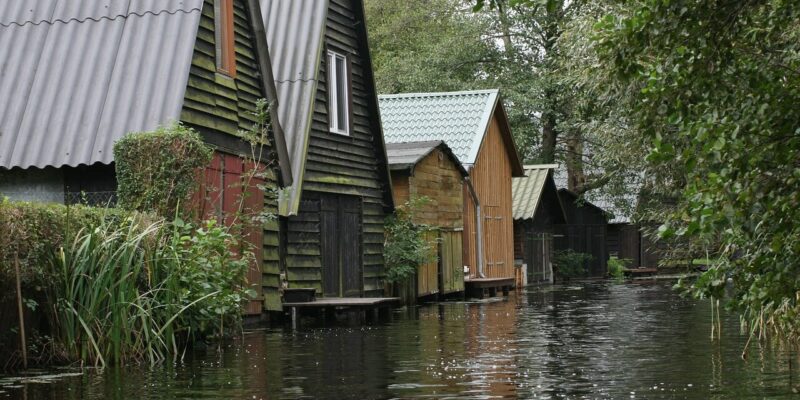Eine Reihe charmanter Holzhäuser mit Satteldächern säumt einen schmalen Kanal, der an die Müritz erinnert, umgeben von üppiger grüner Vegetation. Die Spiegelbilder der Häuser und Bäume spiegeln sich wunderschön im ruhigen Wasser.
